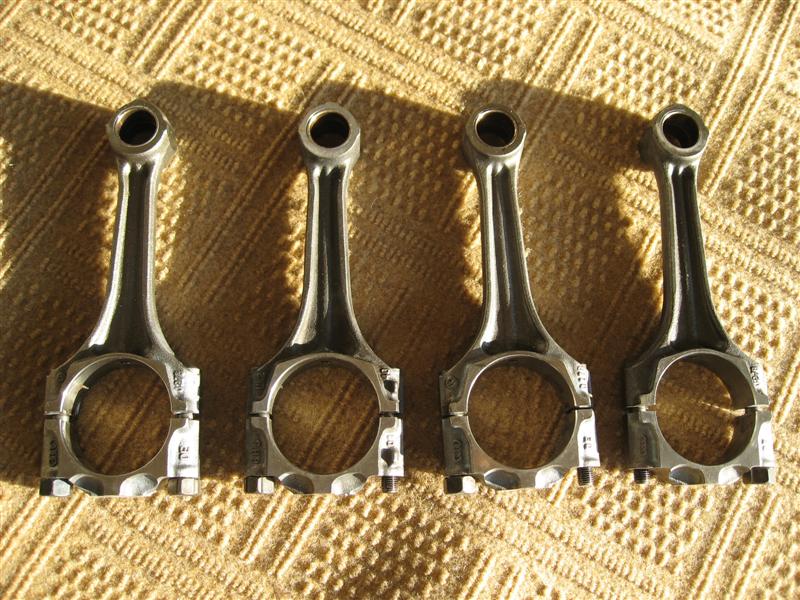 Four polished engine connecting rods laid in a row on a tan blanket in sunlight