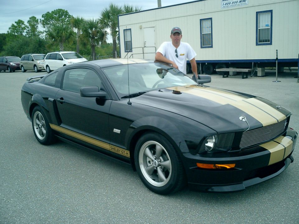 Black Shelby GT-H Mustang coupe with gold stripes, 3/4 front view, owner standing behind it