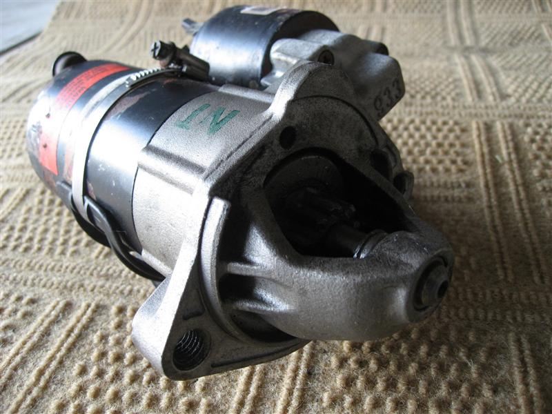Starter motor with metal casing and black cylindrical component, resting on a textured surface