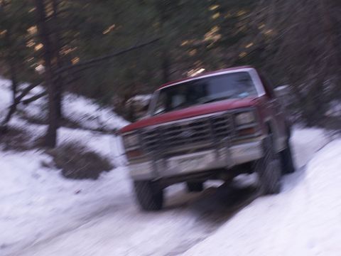 1986 Ford Bronco - A red pickup truck navigates a snowy path in a wooded area.