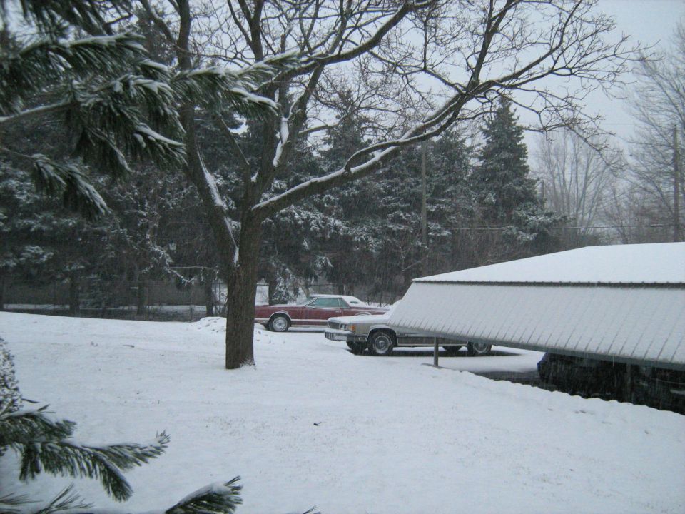 Two cars parked outdoors in falling snow near a barn