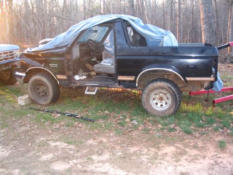 1987 Ford Bronco - A black Ford Bronco sits outdoors with its driver's door open and a tarp covering part of its body.