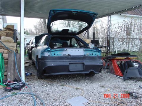 1993 Ford Probe - A car in disrepair is parked under a carport with other vehicle parts nearby.