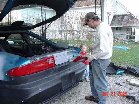 1993 Ford Probe - Man and child working on the rear of a 1993 Ford Probe, with the trunk open and parts removed.