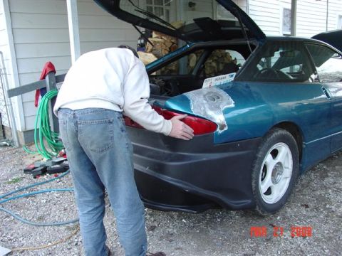 1993 Ford Probe - A person works on the rear of a teal coupe, which has its bumper removed and appears to be undergoing repairs.