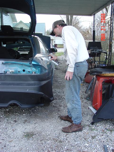 1993 Ford Probe - A person is spray painting the rear bumper of a teal-colored car in an outdoor setting.