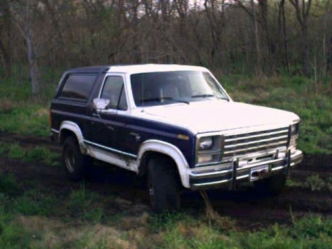 1981 Ford Bronco - Two-tone blue-and-white full-size Ford Bronco with brush guard parked in a muddy grass field with bare trees in the background.