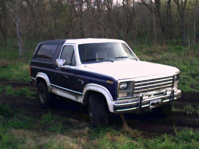 Blue and white full-size Ford Bronco parked in a muddy field with bare trees behind