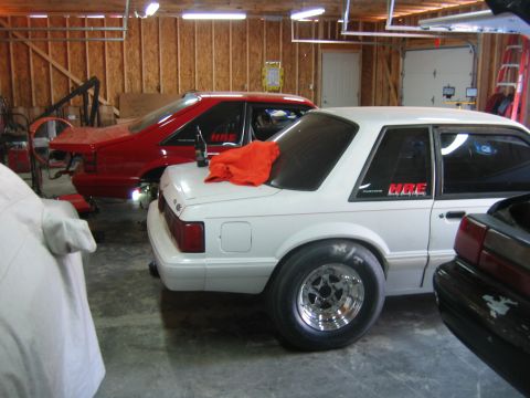 1992 Ford Mustang - Two Ford Mustangs, one white and one red, are parked in a garage. The white Mustang has HRE decals on its window.