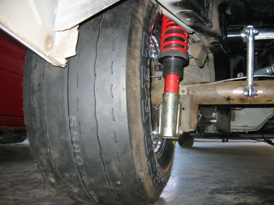 Close-up of a wide drag racing tire with visible sidewall markings and a red coilover shock absorber assembly