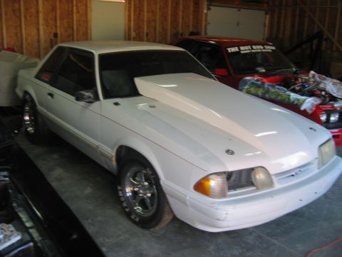 1992 Ford Mustang - A white 1992 Ford Mustang coupe with a prominent hood scoop is parked in a garage, with another vehicle visible behind it.