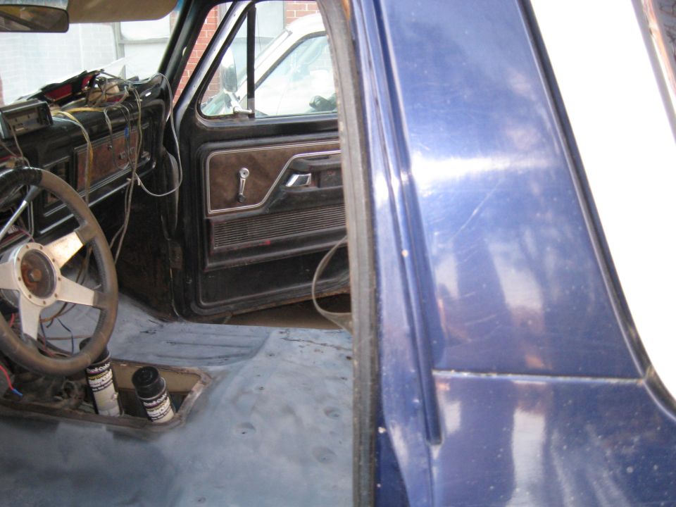 Interior of a truck with exposed floor, aftermarket steering wheel, and door panel visible. Another vehicle is seen through the window