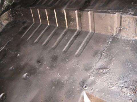 1978 Ford Bronco - The interior floor of a vehicle shows a dark undercoating with distinct ribbed metal sections and some circular indentations.