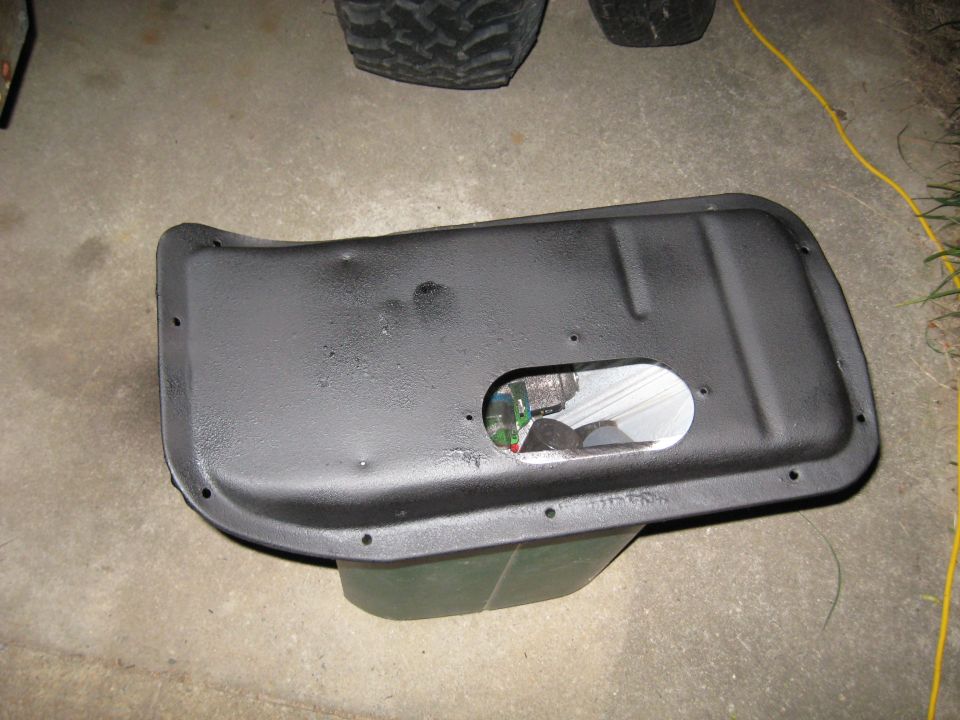 Black metal part with oval cutout revealing electronics, resting on a green object. Tire treads visible in background
