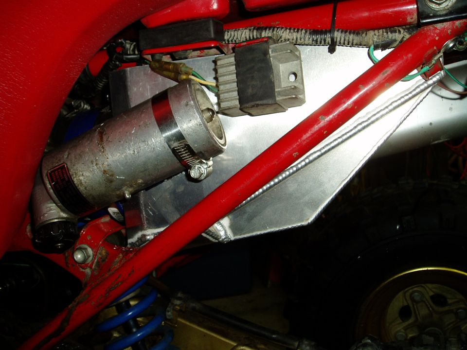 Close-up of a silver cylinder with red bands, a black voltage regulator, and red frame tubes on a red vehicle