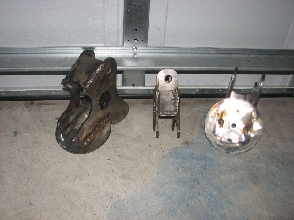 Three welded steel suspension brackets on a garage floor in front of a white door.