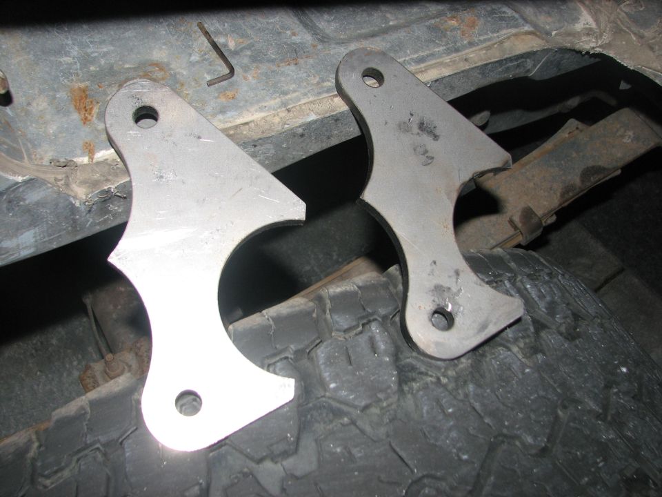Two raw steel fabricated brackets with bolt holes resting on a tire under a rusty vehicle underbody