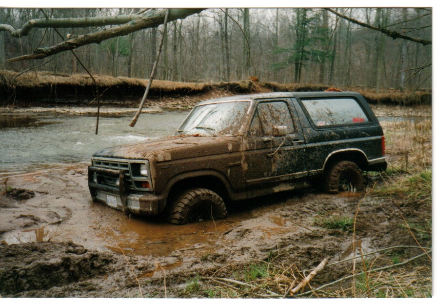 Mud-caked dark Ford Bronco stuck in deep mud next to a creek in a bare winter forest