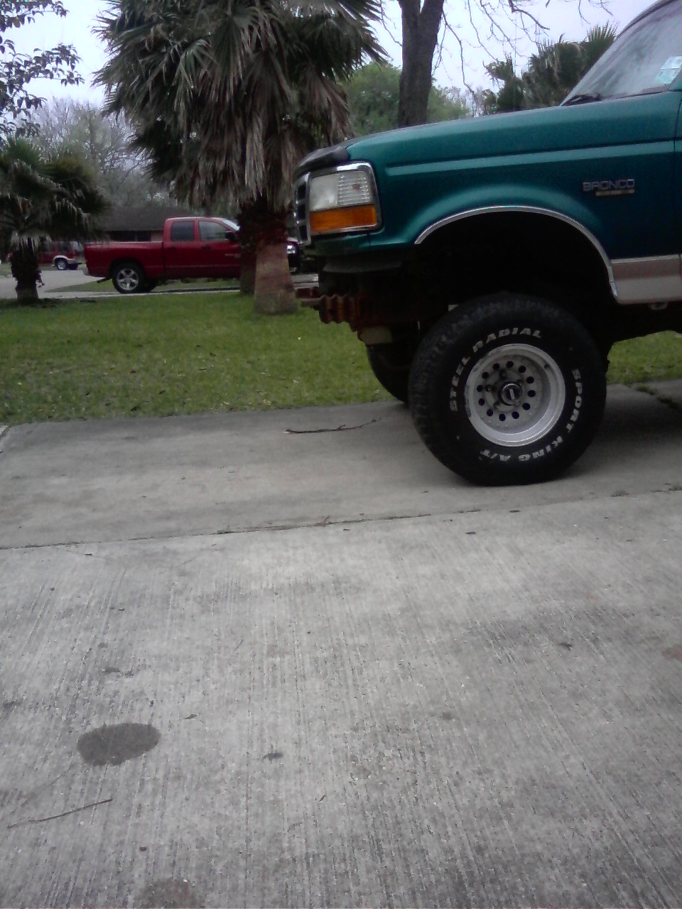Green Ford Bronco, front quarter panel and wheel visible. Red pickup truck in background