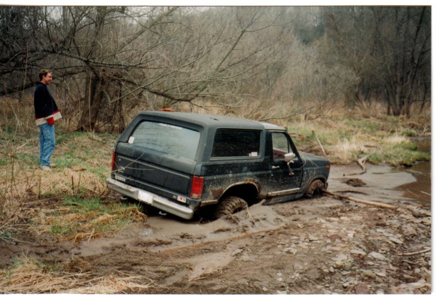 Dark Ford Bronco stuck axle-deep in mud by a creek, man watching from grassy bank