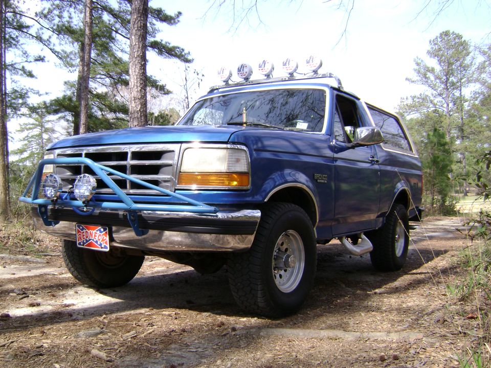 Blue Ford Bronco SUV with brush guard, roof lights, and off-road tires on a wooded dirt trail