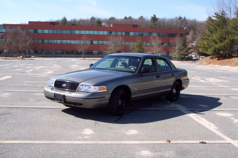 2001 Ford Crown Victoria - A gray 2001 Ford Crown Victoria is parked in a lot, with a large red brick building behind it.
