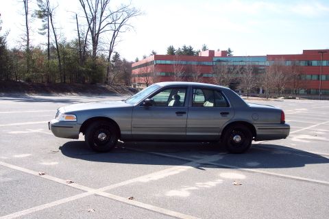 2001 Ford Crown Victoria - A gray 2001 Ford Crown Victoria sedan is parked in a large, empty parking lot with a modern building in the background.