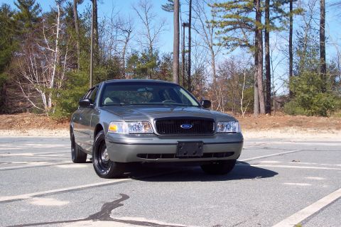 2001 Ford Crown Victoria - Front three-quarter view of a light sage-green Ford Crown Victoria police-package sedan parked in a sunny lot, with a blacked-out grille, n…