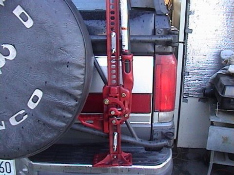 1990 Ford Bronco - A red hi-lift jack is mounted to the rear of a 1987 Ford Bronco, with a spare tire cover on the left.