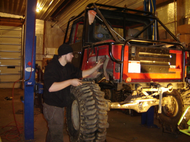 Red off-road vehicle with roll cage and large tires on a lift, being worked on by a person