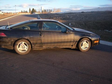 1990 Ford Probe - A black 1990 Ford Probe is seen from the side, parked on an asphalt surface under a cloudy sky.