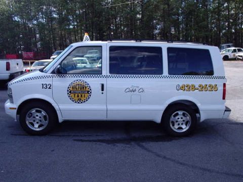 1996 Ford Crown Victoria - A white taxi van is parked on asphalt with trees in the background.