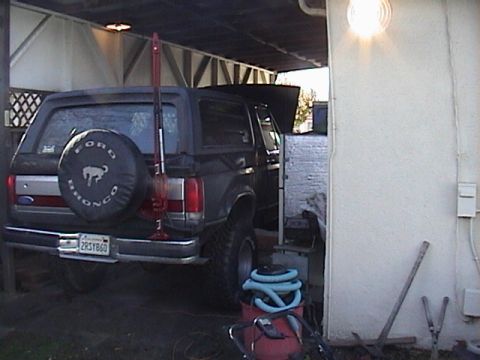 1990 Ford Bronco - A black Ford Bronco is parked under a cover, with a Hi-Lift jack attached to the rear.