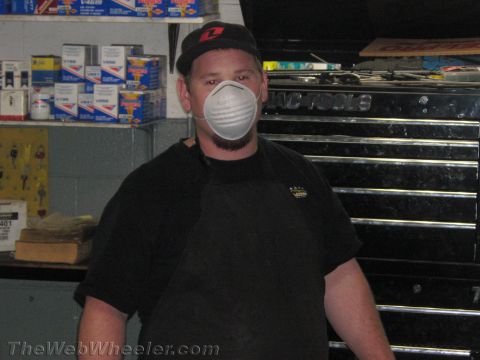 1987 Suzuki Samurai - A man wearing a dust mask and apron stands in a workshop, with shelves of parts and a tool chest visible in the background.