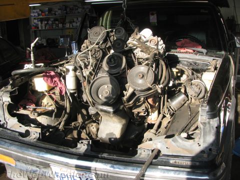 1987 Suzuki Samurai - A V-configuration engine with its pulleys and accessories still attached hangs from a chain hoist over the stripped engine bay of a dark tr…