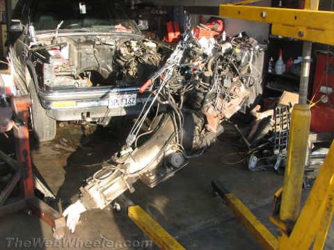 1987 Suzuki Samurai - An engine and automatic transmission assembly hangs from a yellow engine hoist/cherry picker in a garage, suspended in front of a dark SUV/…