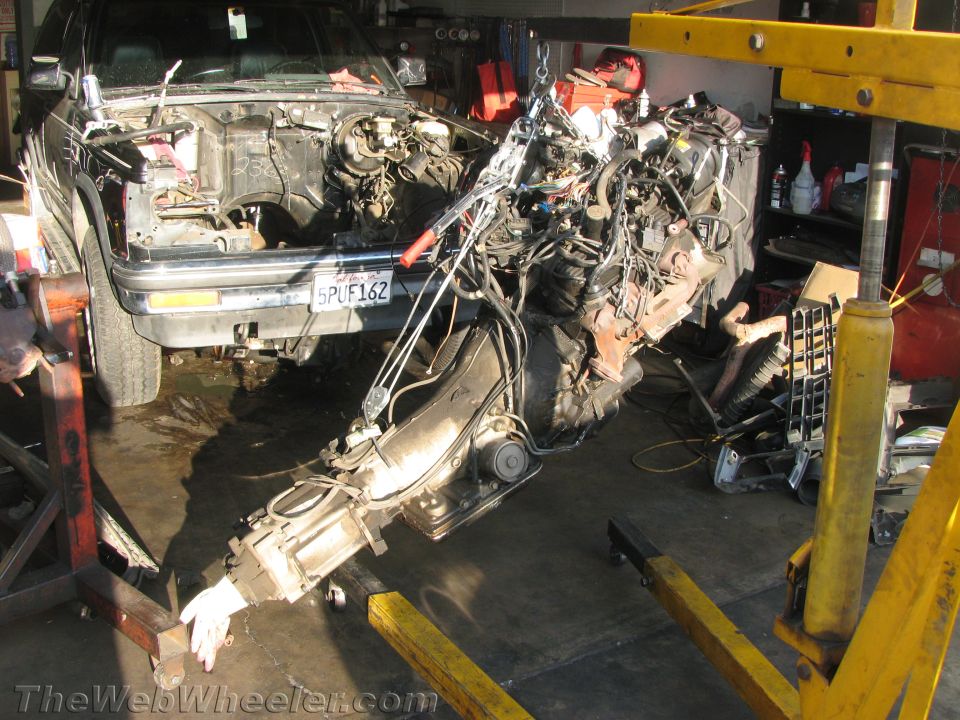 Engine and transmission hanging from yellow hoist in garage near a dark vehicle with removed hood