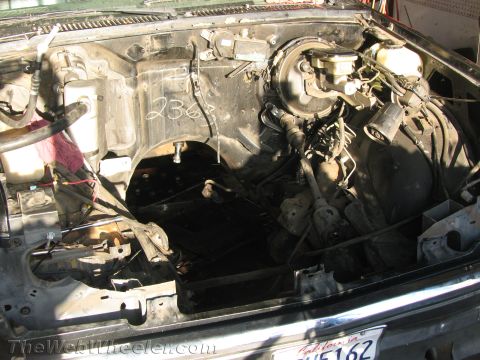 1987 Suzuki Samurai - Overhead view into a disassembled engine bay with the engine, radiator and battery removed, leaving the bare inner fenders, wiring harness,…