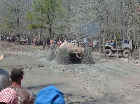 1990 Ford F-150 - Multiple off-road vehicles navigate a muddy track during an event, with onlookers present.