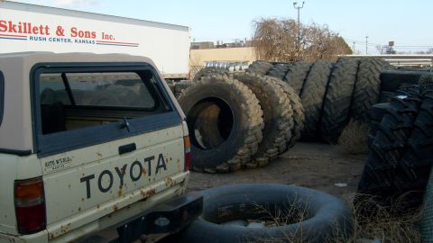 1995 Ford C-350 (Centurion Conversions) - A Toyota pickup truck is parked next to a stack of large tires and a semi-trailer, suggesting a commercial or industrial setting.