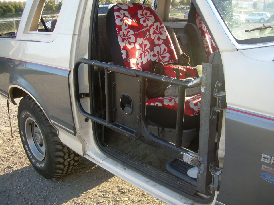 White Ford Bronco interior with red floral seat covers and a black roll cage around the driver's seat. The door is open