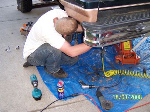 1991 Ford Bronco - A person works on the rear bumper of a dark green SUV, surrounded by tools and a jack on a blue tarp.