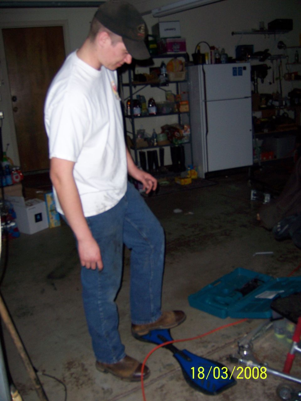 Person in a garage with shelves, a refrigerator, and a blue device on the floor with a date stamp