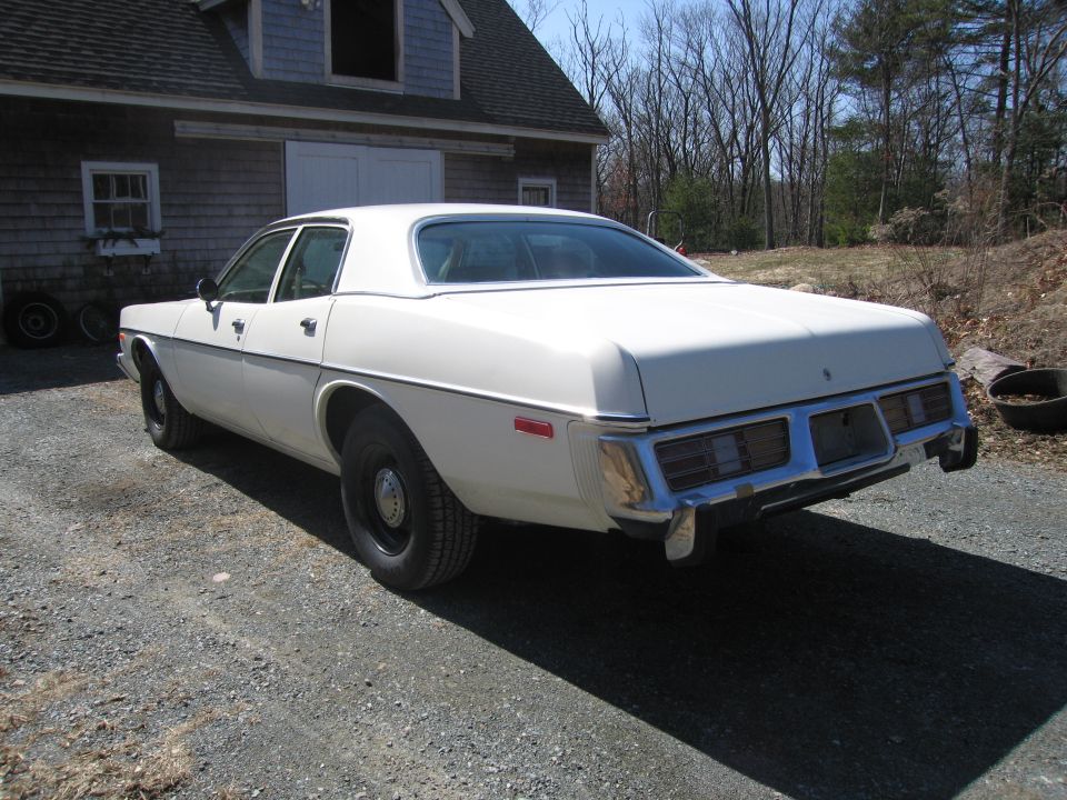 White 1977 sedan, rear 3/4 view, parked on gravel in front of a barn