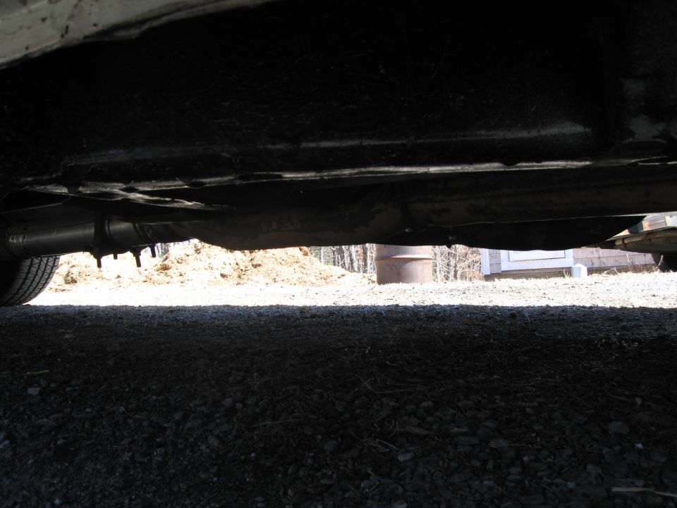 Underside view of a vehicle, showing the exhaust pipe and frame. Gravel and a dirt pile are visible in the background