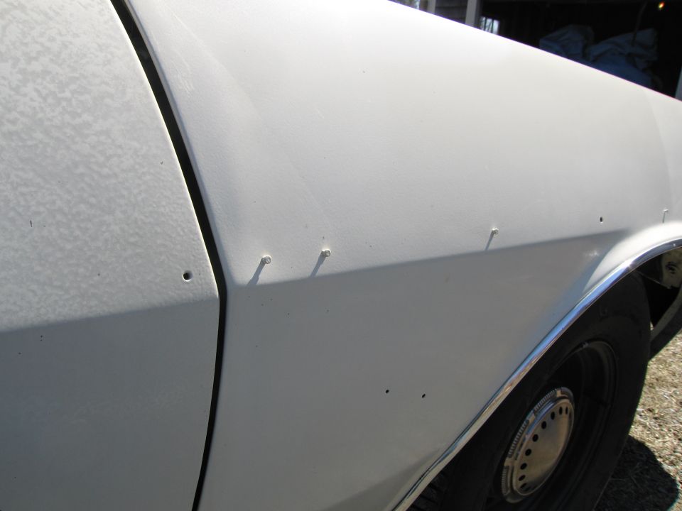 White car fender with visible screw holes and a chrome trim piece above the wheel