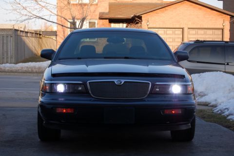 1985 Mercury Grand Marquis - The front end of a Mercury Grand Marquis is shown with its headlights on, parked in front of a house.
