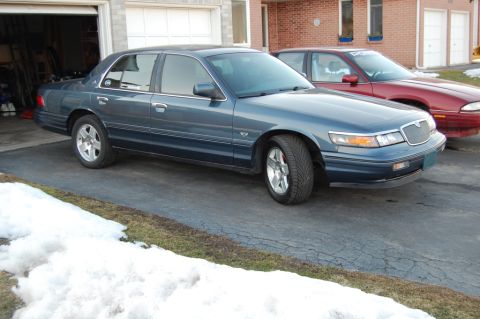 1985 Mercury Grand Marquis - A blue sedan is parked in a driveway with snow on the ground, next to a red car.