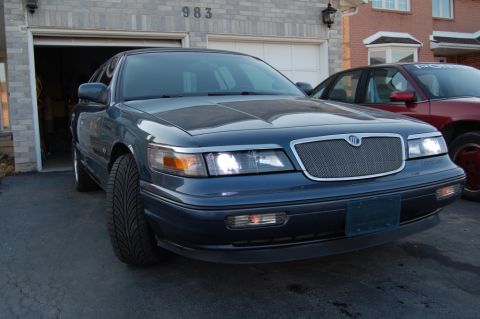 1985 Mercury Grand Marquis - A dark blue sedan is parked in front of a garage, with another car visible to its right.