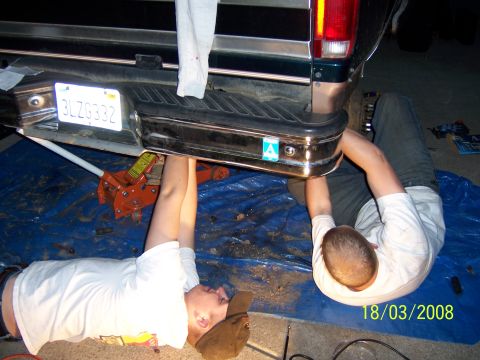 1991 Ford Bronco - Two individuals work on the rear bumper of a dark green Ford Bronco, supported by a jack.